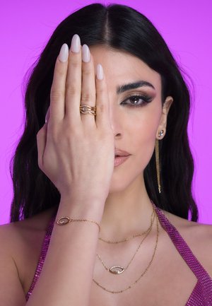 Goldschmuck mit geschichteten Halsketten, die Augen-Anhänger tragen, gestapelten Ringen und langen hängenden Ohrringen, auf einem lila Hintergrund präsentiert.
