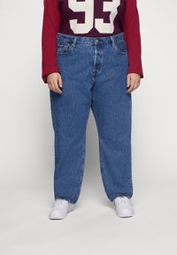 Vaqueros de mezclilla azul con un ajuste relajado, cinco bolsillos y cierre de botón. Combinados con una camisa de manga larga color burdeos y zapatillas blancas.