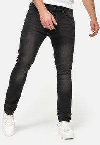 Zwarte denim jeans met een slim fit, vijf zakken en contrasterende stiksel. Gecombineerd met witte sneakers op een effen achtergrond.