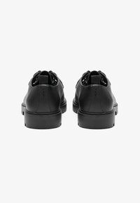 Zapatos de cuero negro con una textura suave, puntera redondeada y suela de plataforma baja. Presenta una lengüeta en la parte posterior para ponerlos y quitarlos con facilidad.