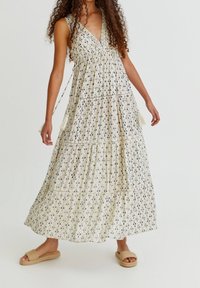 Vestido maxi en crema con un patrón navy, escote en V profundo, cuerpo fruncido y falda en capas. Presenta detalles de borlas y se combina con sandalias planas.