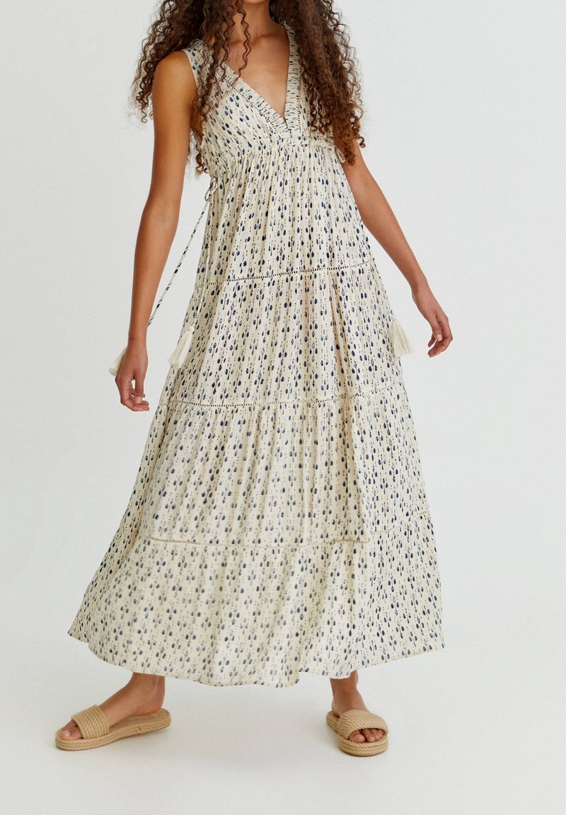 Vestido maxi en crema con un patrón navy, escote en V profundo, cuerpo fruncido y falda en capas. Presenta detalles de borlas y se combina con sandalias planas.