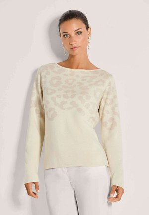 Vrouw met naar achteren gekamd haar, draagend gouden abstracte oorbellen en een beige trui met een subtiel luipaardpatroon, staand tegen een lichte achtergrond.