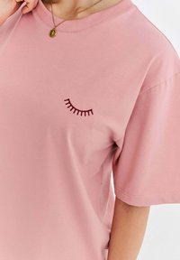 Růžové bavlněné tričko s krátkým rukávem a vyšitým detailem řas na levé přední části. Hladká textura a kulatý výstřih.