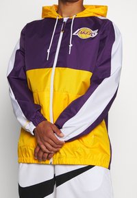 Lilla og gul vindjakke med hætte, udstyret med elastiske manchetter og lynlås foran. Viser et Los Angeles Lakers-logo på brystet.