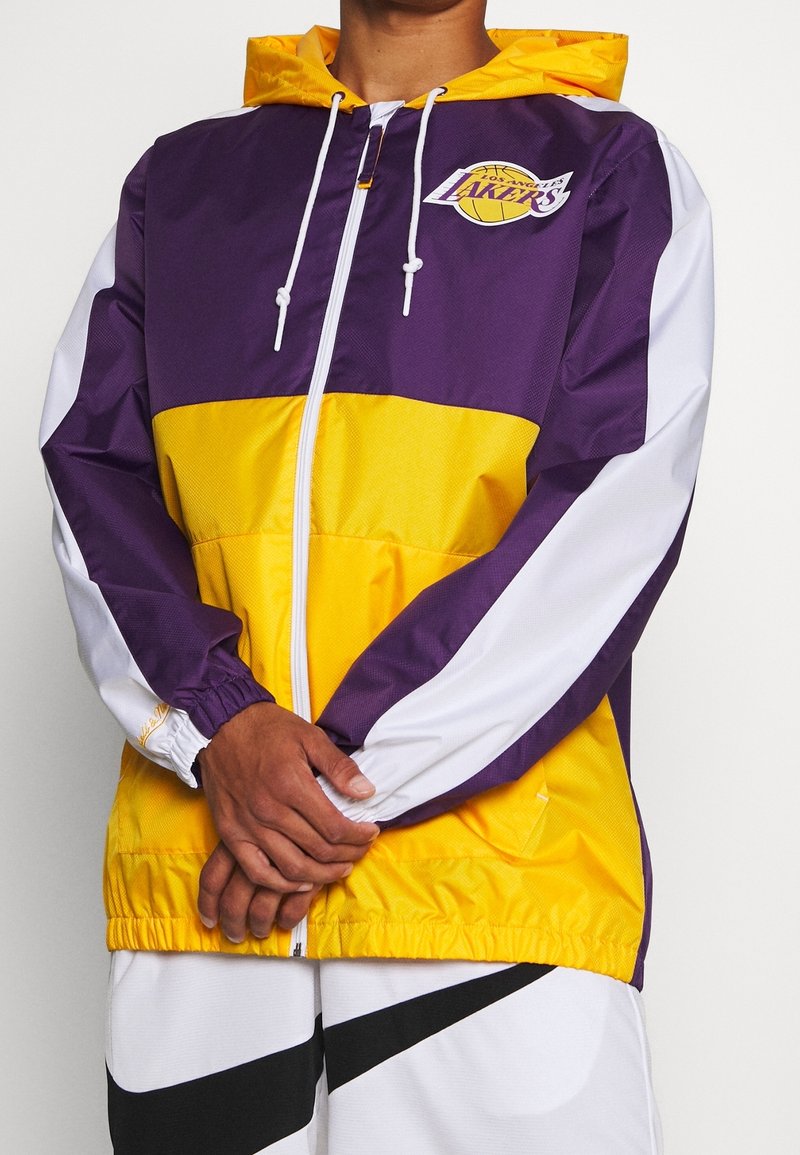 Lilla og gul vindjakke med hætte, udstyret med elastiske manchetter og lynlås foran. Viser et Los Angeles Lakers-logo på brystet.