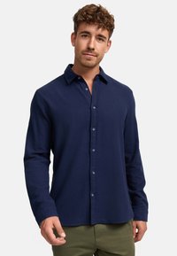 Camisa de botones de manga larga en navy, con un tejido texturizado, cuello ajustado y bolsillos frontales, que presenta botones azules y un logo sutil.