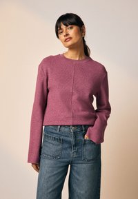 Pull-over en tissu doux et texturé de couleur rose profond, avec un col rond, des manches longues, et une coupe légèrement courte. Associé à un jean bleu taille haute.