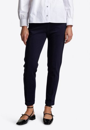 La personne porte un pantalon noir ajusté roulé aux chevilles, une chemise blanche boutonnée et des escarpins noirs en cuir verni à talons bas avec des brides autour de la cheville.