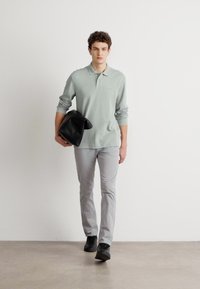 Light green long-sleeve polo, textured fabric, gray slim-fit trousers, and black leather bag; styled with black sneakers. Simple backdrop.