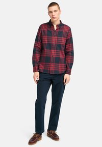 Red and navy plaid flannel shirt with long sleeves, button-up front, and a single chest pocket, paired with navy trousers and brown shoes.