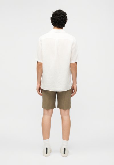 Άνδρας με σγουρά μαλλιά, που φοράει ένα λευκό κοντομάνικο πουκάμισο, χακί πράσινα σορτς, λευκές κάλτσες και λευκά αθλητικά παπούτσια, στέκεται με την πλάτη γυρισμένη.