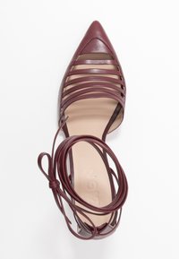 Scarpa in pelle bordeaux con punta affusolata, dotata di tagli orizzontali e un dettaglio di lacci avvolgenti. Texture liscia e suola interna piatta.
