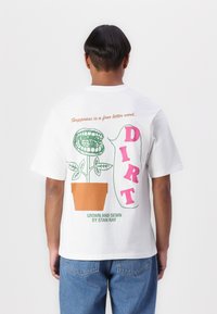 White short-sleeve t-shirt featuring a graphic of a plant in a pot with text "Happiness is a four letter word" and "DIRT" in pink.