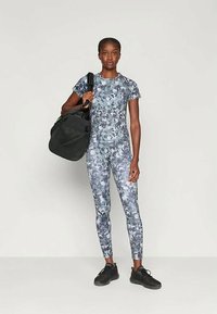 Naine kannab halli-sinist kamuflaaži sportlikku riietust, hoides ühte õlavarre peal musta spordikotti, seistes ühetoonilise heleda tausta vastas.