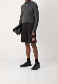 Polo gris à manches longues avec une fermeture éclair, associé à un short noir arborant un patch logo, et des chaussures noires à lacets avec une semelle épaisse.