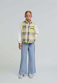 Chaleco de forro polar con un patrón de cuadros en crema, marrón y púrpura, con un bolsillo y cremallera en verde neón. Jeans de mezclilla de pierna ancha con rayas laterales.