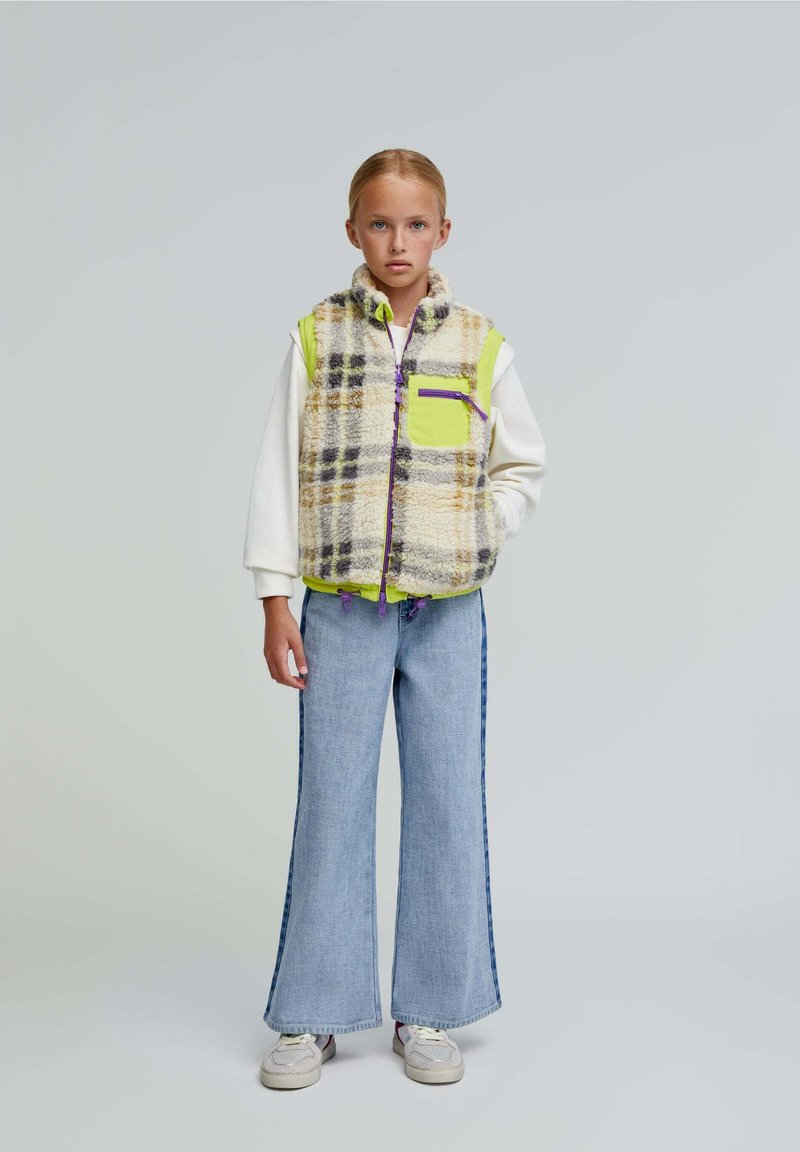 Chaleco de forro polar con un patrón de cuadros en crema, marrón y púrpura, con un bolsillo y cremallera en verde neón. Jeans de mezclilla de pierna ancha con rayas laterales.