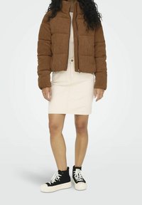 Bruine corduroy pufferjack met hoge kraag, voorzijde met rits; gecombineerd met een lichtbeige rok tot de knie en zwarte high-top sneakers.