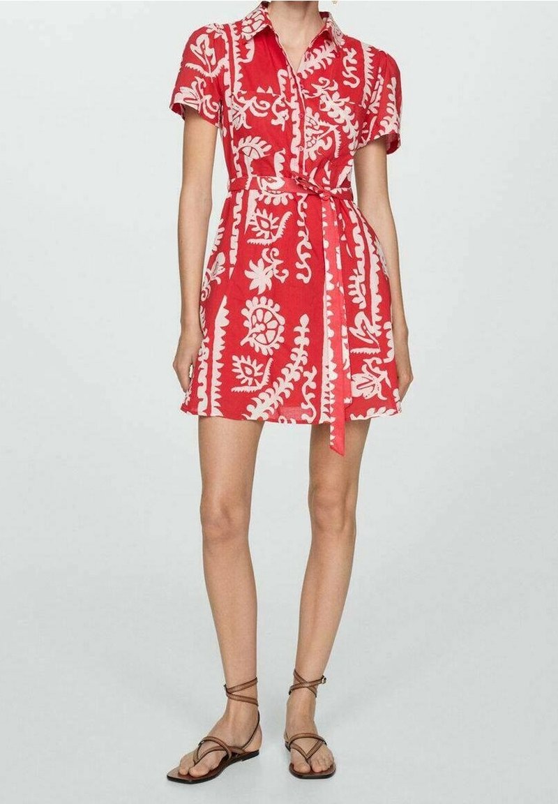 Femme portant une robe rouge à manches courtes avec des motifs floraux blancs, une ceinture à la taille, et des sandales plates marron à brides, debout sur un fond uni.