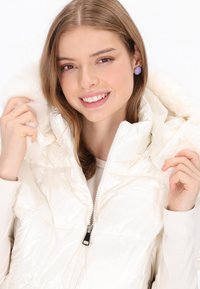 Biała pikowana kurtka puchowa z błyszczącą, przeszywaną powierzchnią, wyposażona w kaptur obszyty futrem oraz czarny zamek błyskawiczny. Modelka ma pod spodem lekką sweter.