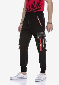 Schwarze Cargo-Hosen mit mehreren Taschen, roten Akzenten und verstellbaren Bündchen. Hergestellt aus strapazierfähigem Stoff mit glatter Textur und Reißverschlussdetails.