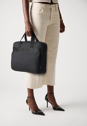 Black laptop bag with dual handles, front zipper pocket, and brand logo, made from durable material, held by a person in light-colored pants.