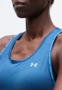 Κοντινό πλάνο ενός ατόμου που φοράει μπλε αθλητικό τοπ της Under Armour με λουράκια με λογότυπο και ορατή περιοχή άνω στήθους και λαιμού.