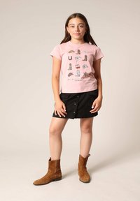 Lichtroze t-shirt met grafische prints, zwarte minischort met knopen aan de voorkant, en bruine suède enkellaarzen. Eenvoudige en casual outfit.