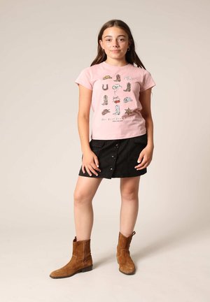 Lichtroze t-shirt met grafische prints, zwarte minischort met knopen aan de voorkant, en bruine suède enkellaarzen. Eenvoudige en casual outfit.