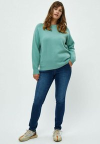 Maglione oversize verde chiaro con polsini a costine, abbinato a jeans skinny blu scuro e sneakers in tonalità neutre. Outfit semplice e informale.