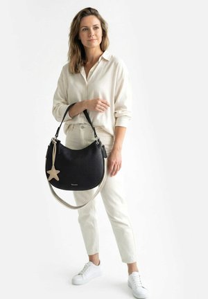 Vrouw in witte blouse en broek, met een grote zwarte handtas met een beige stervormige hanger, draagt witte sneakers, staand op een witte achtergrond.