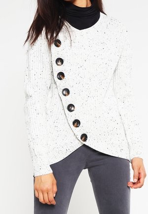 Kobieta ubrana w biały dzianinowy sweter w kropki z dużymi czarnymi ukośnymi guzikami, na czarnym golfie i szarych spodniach.