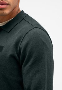 Dunkel gestricktes Polohemd mit Kragen, strukturiertem Stoff und einem dezenten Badge am Ärmel. Nahaufnahme, die die details der Schulternaht zeigt.