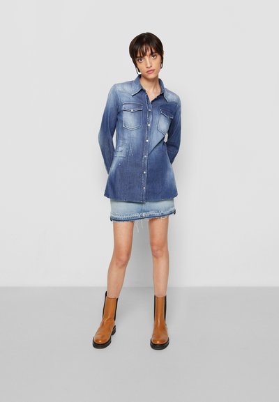 Camisa de ganga com fecho de botão, dois bolsos no peito e um tom azul desgastado. Combinada com uma saia de ganga desgastada e botas de tornozelo castanhas.