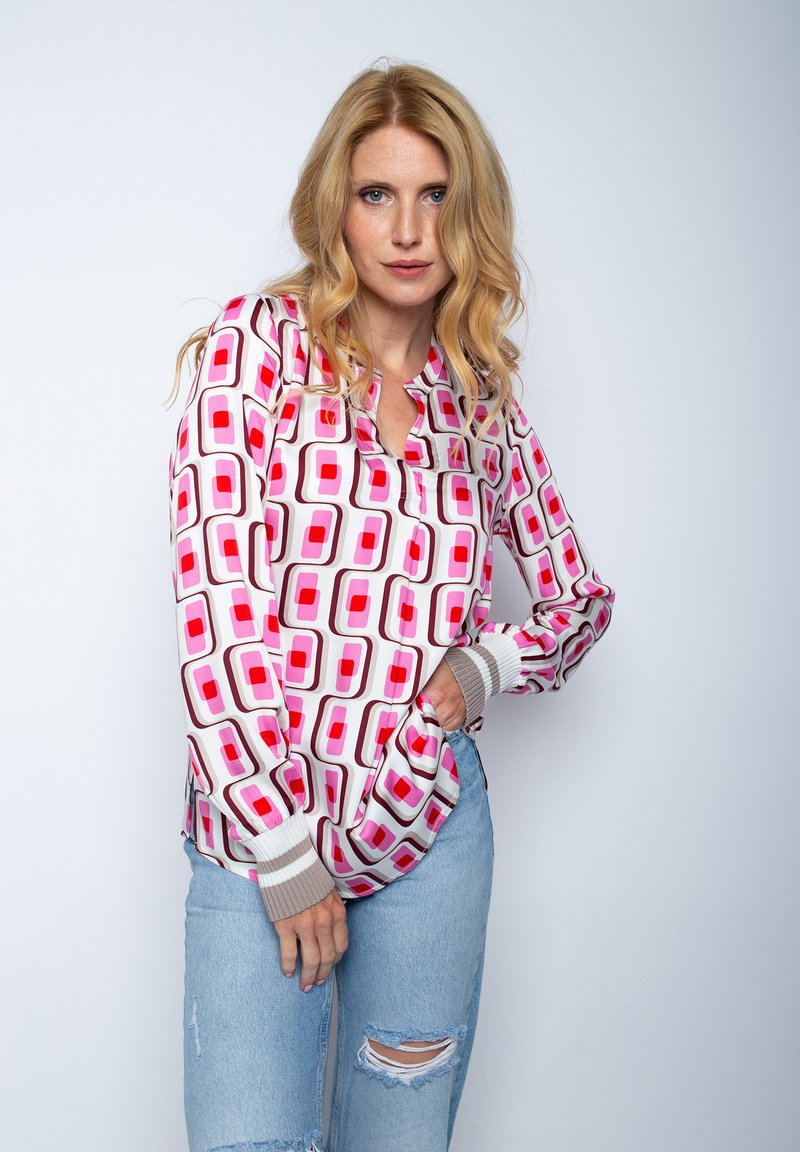 Bunte Bluse mit auffälligem geometrischem Muster in Pink und Rot auf weißem Hintergrund. Lange Ärmel mit grauen Bündchen. Kombiniert mit hellblauen, verwaschenen Jeans.