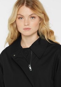 Blonde Frau mit welligem Haar trägt eine schwarze Zip-Jacke mit Kragen und blickt auf einem schlichten weißen Hintergrund geradeaus.