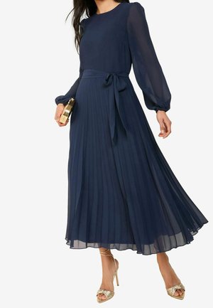 Femme portant une robe midi plissée bleu marine à manches longues avec une ceinture à la taille, tenant une pochette dorée et portant des sandales à talons hauts dorées.