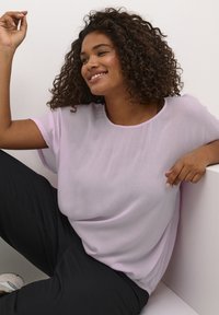Helllila, lockere Bluse mit rundem Halsausschnitt und kurzen Ärmeln, aus weichem, durchscheinendem Stoff, kombiniert mit schwarzen Hosen.