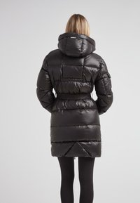 Mujer vestida con un largo abrigo de plumas negro con capucha, de pie con las manos en los bolsillos, vista desde atrás contra un fondo gris liso.