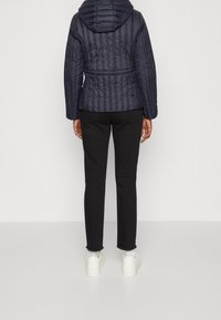 Marinblå, vadderad puffjacka med huva, figursydd i midjan, och svarta jeans. Jeans med slitna kanter; vita sneakers synliga.