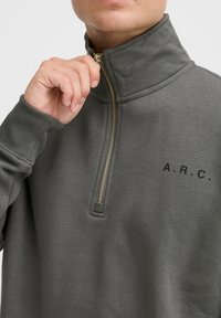 Sudadera gris con cuello alto, cremallera de cuarto y cremallera dorada. Presenta un logo impreso "A.R.C." en el lado izquierdo. Material de algodón con una textura suave.