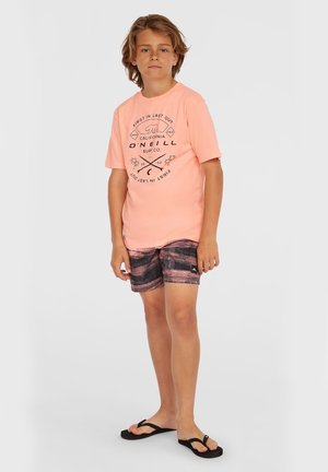 Camiseta de color rosa claro con texto gráfico negro, combinada con pantalones cortos de baño a rayas multicolores. Las chanclas negras completan el atuendo.