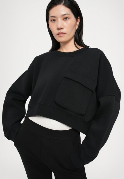 Black cropped sweatshirt with a front pocket, ribbed crew neckline, and textured logo. Constructed from soft, stretchy fabric, paired with black pants.