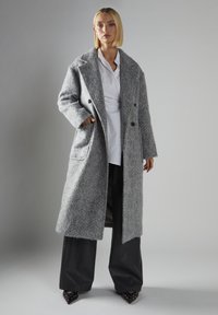 Langer, grauer Wollmischmantel mit strukturierter Oberfläche, großen Revers und Seitentaschen. Kombiniert mit weiten schwarzen Hosen und Absatzschuhen.