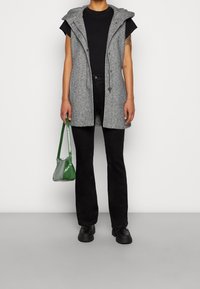 Szara bezrękawnikowa kamizelka z kapturem, zapinana na guziki i ze sznurkiem, w komplecie z czarną koszulką, czarnymi dżinsami oraz zieloną torebką o fakturze.