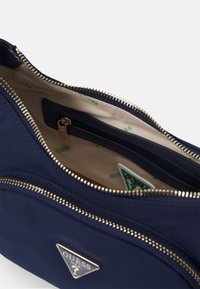 Sac Guess bleu marine avec deux compartiments zippés, doublure intérieure beige imprimée de logos Guess verts et une petite poche intérieure zippée.