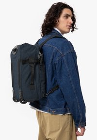 Tummansininen kangaskassi pyörillä, useilla hihnoilla ja yläkahvalla. Ominaisuuksiin kuuluu teksturoitu pinta ja kontrastiväriset mustat metalliosat.