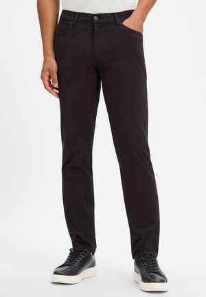 Mann trägt schwarze Slim-Fit-Hose mit schwarzen Ledersneakern und weißen Sohlen, eine Hand in der Tasche, steht vor weißem Hintergrund.