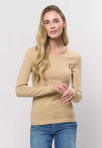 Beige långärmad topp med ribbad textur och en liten broderad detalj på bröstet. Kombinerad med blå denimjeans.